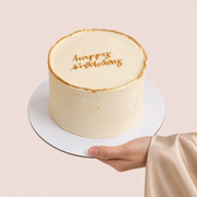 A round 1kg TWIGS Gold celebration cake with gold trim and a "Happy Birthday" message piped in gold icing, held on a white cake stand.