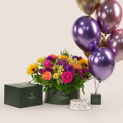 Festive grande round flower box with sunflowers, roses, alstroemerias, chrysanthemums, and wax flowers, paired with a 1kg birthday cake and six chrome balloons in TWIGS beige or green packaging, available for same day flower delivery in Dubai when you buy flowers online.