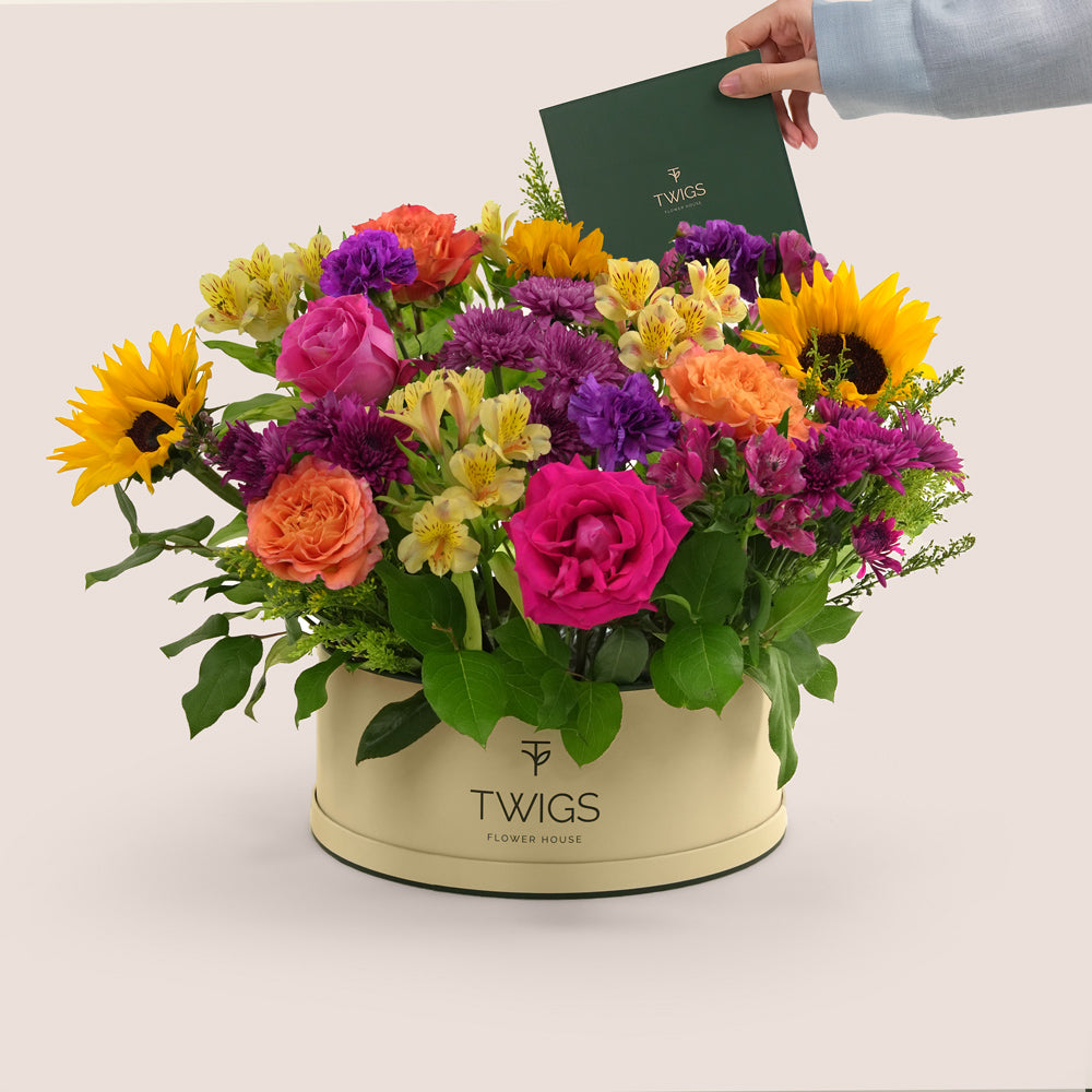 Festive grande round flower box with sunflowers, roses, alstroemerias, chrysanthemums, and wax flowers in TWIGS beige or green packaging, available for same day flower delivery in Dubai when you buy flowers online.