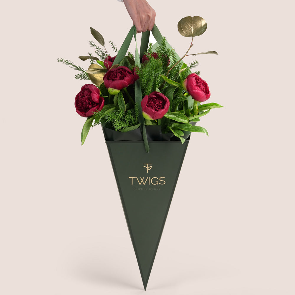 beautiful Christmas bouquet of deep red peonies, lush green pine needles, and gold-painted foliage, presented in a distinctive green TWIGS Flower House cone carrier bag 