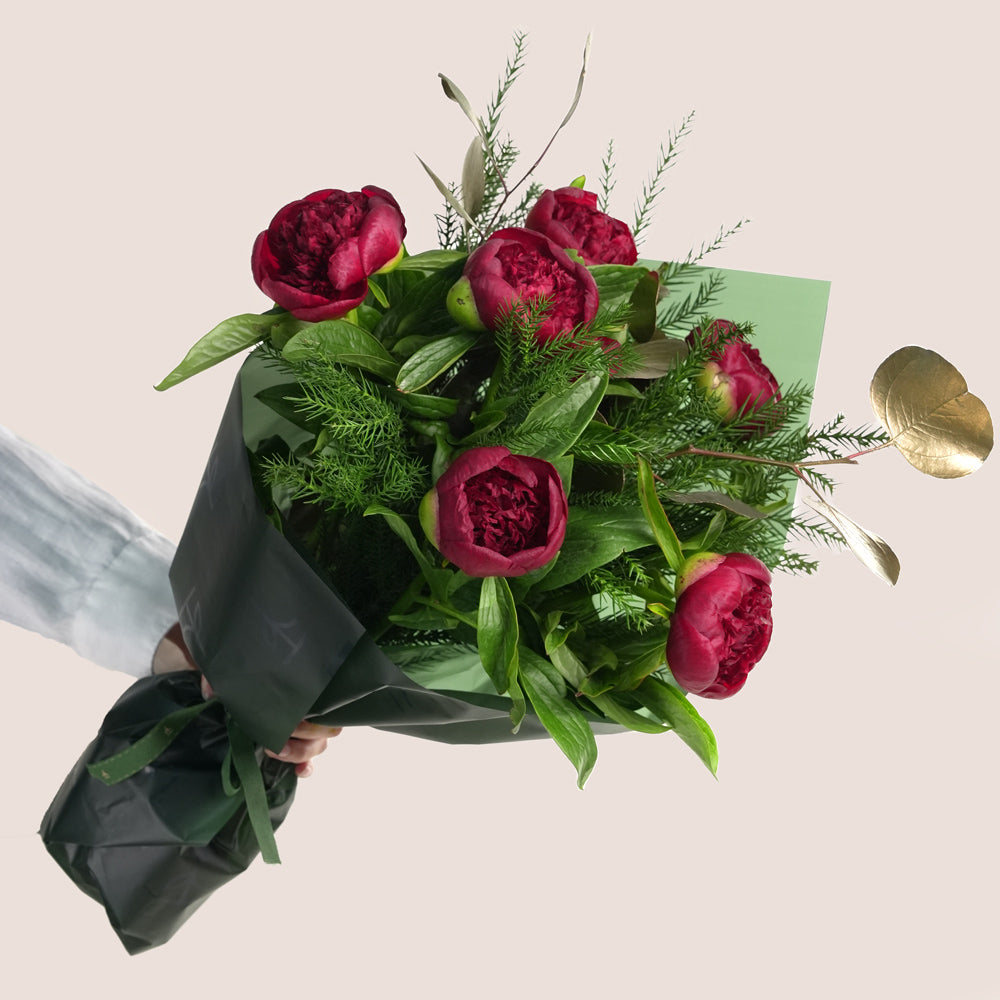 beautiful Christmas bouquet of deep red peonies, lush green pine needles, and gold-painted foliage, presented in a distinctive green TWIGS Flower House cone carrier bag.