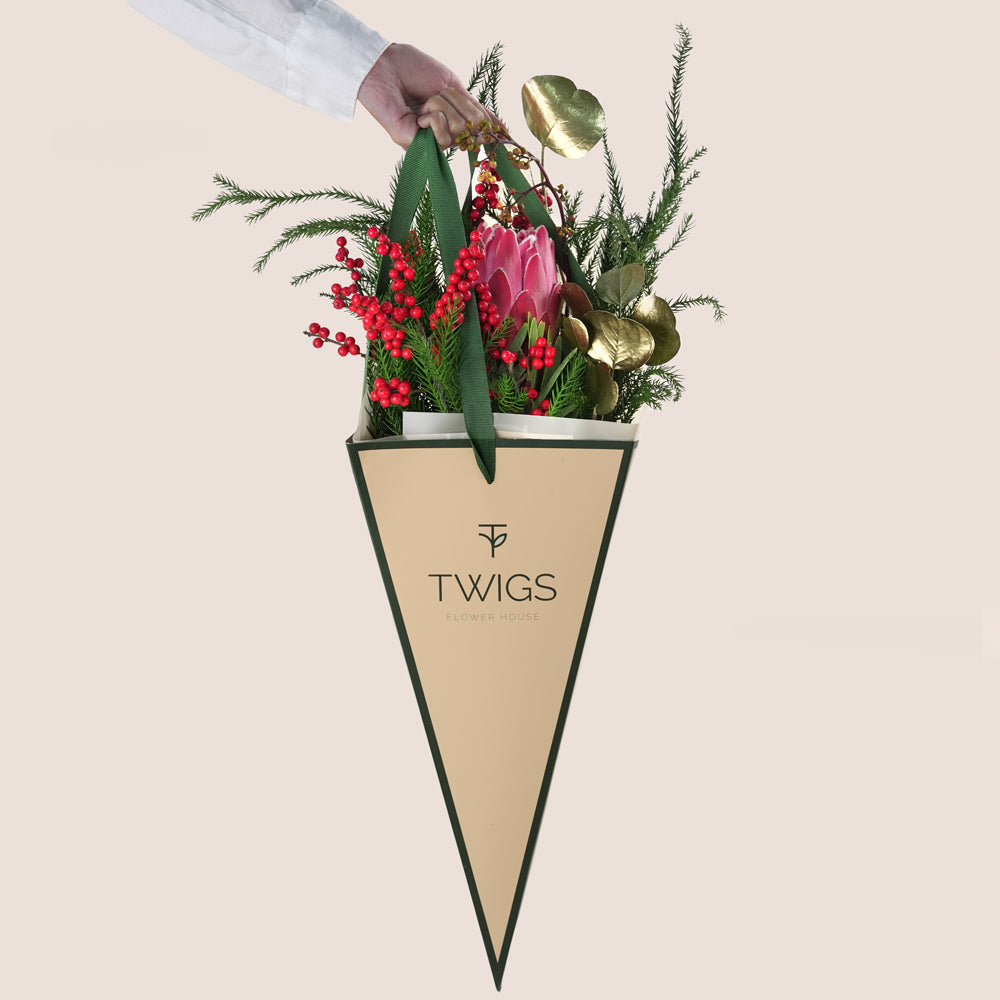 Christmas flower bouquet featuring a central pink King Protea, vibrant red hypericum berries, evergreen foliage, and gold-painted leaves, all in a beige and green TWIGS cone carrier bag