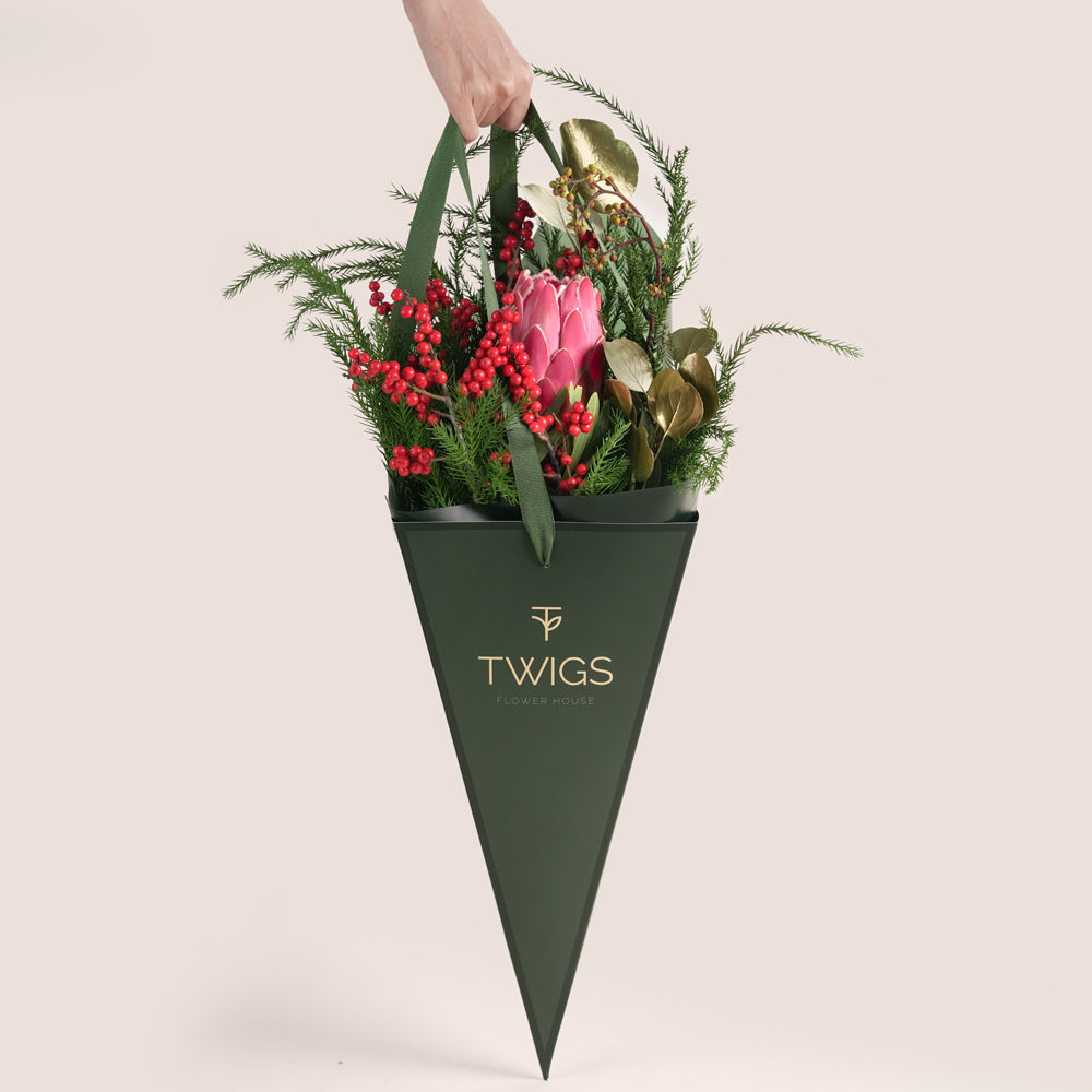 Christmas flower bouquet featuring a central pink King Protea, vibrant red hypericum berries, evergreen foliage, and gold-painted leaves, all in a beige and green TWIGS cone carrier bag