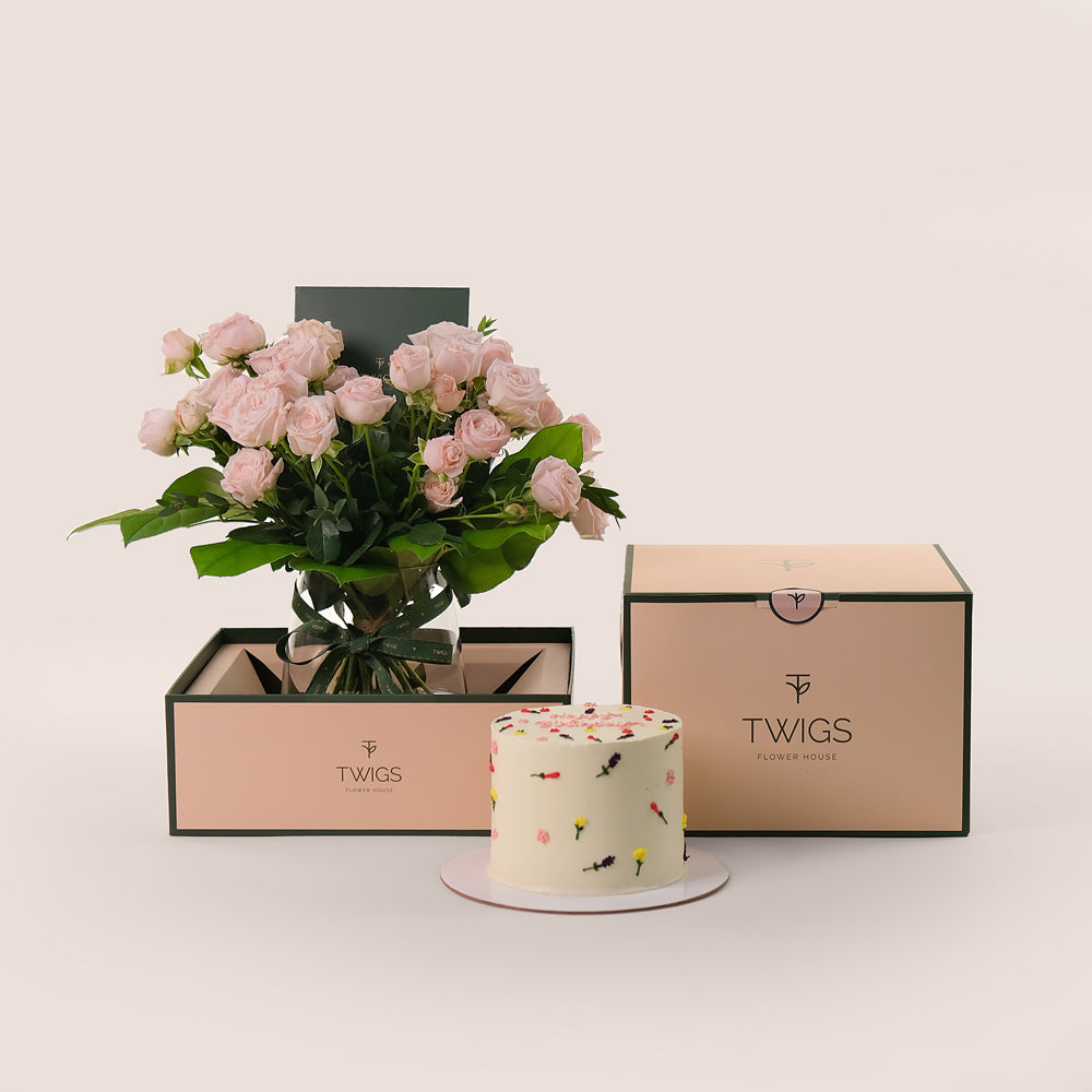 A bouquet of pink roses in a petite glass vase alongside a birthday cake with floral icing decorations and a 'Happy Birthday' message, presented in front of a TWIGS' carry box.