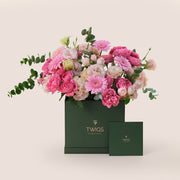 A luxurious floral arrangement featuring fuchsia cerise Juliet roses, pink gerberas, light pink bombastic spray roses, and pastel lisianthus in a square-shaped flower box, with the TWIGS 