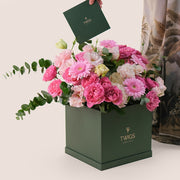 A luxurious floral arrangement featuring fuchsia cerise Juliet roses, pink gerberas, light pink bombastic spray roses, and pastel lisianthus in a square-shaped flower box, with the TWIGS 