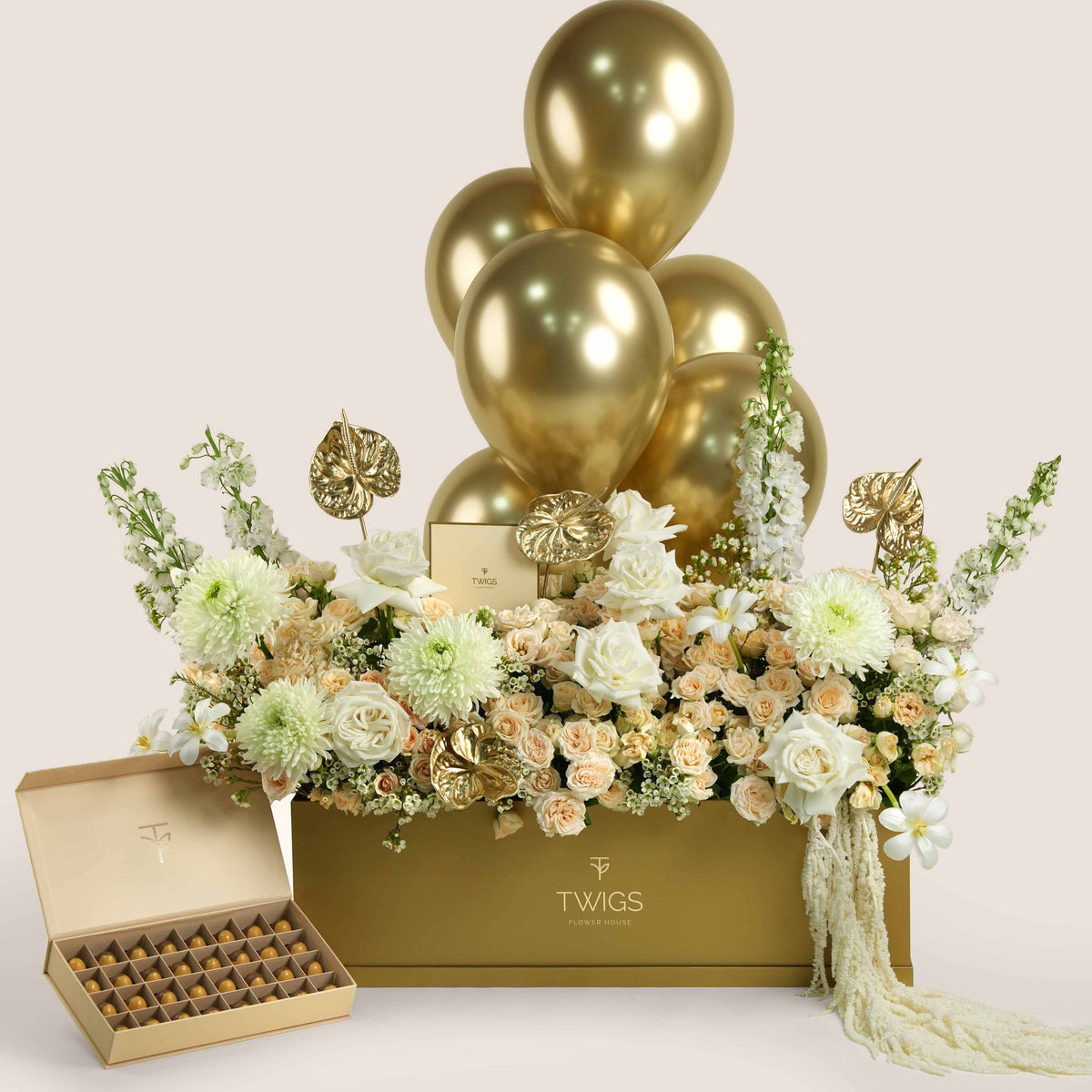Lumina Royale, a luxury TWIGS Gold Garden box arrangement of premium roses, chrysanthemums, delphiniums, and gold anthurium leaves, paired with premium bonbons and six gold chrome balloons.