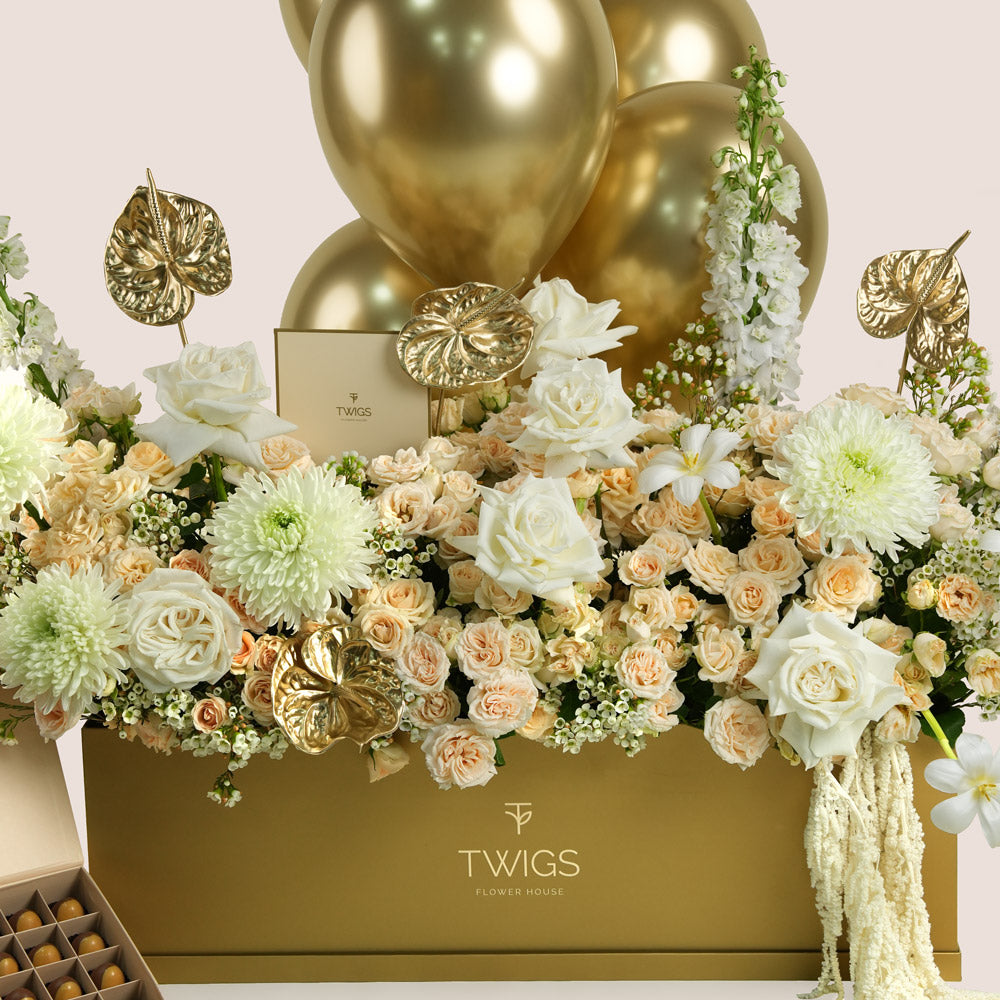 Lumina Royale, a luxury TWIGS Gold Garden box arrangement of premium roses, chrysanthemums, delphiniums, and gold anthurium leaves, paired with premium bonbons and six gold chrome balloons.