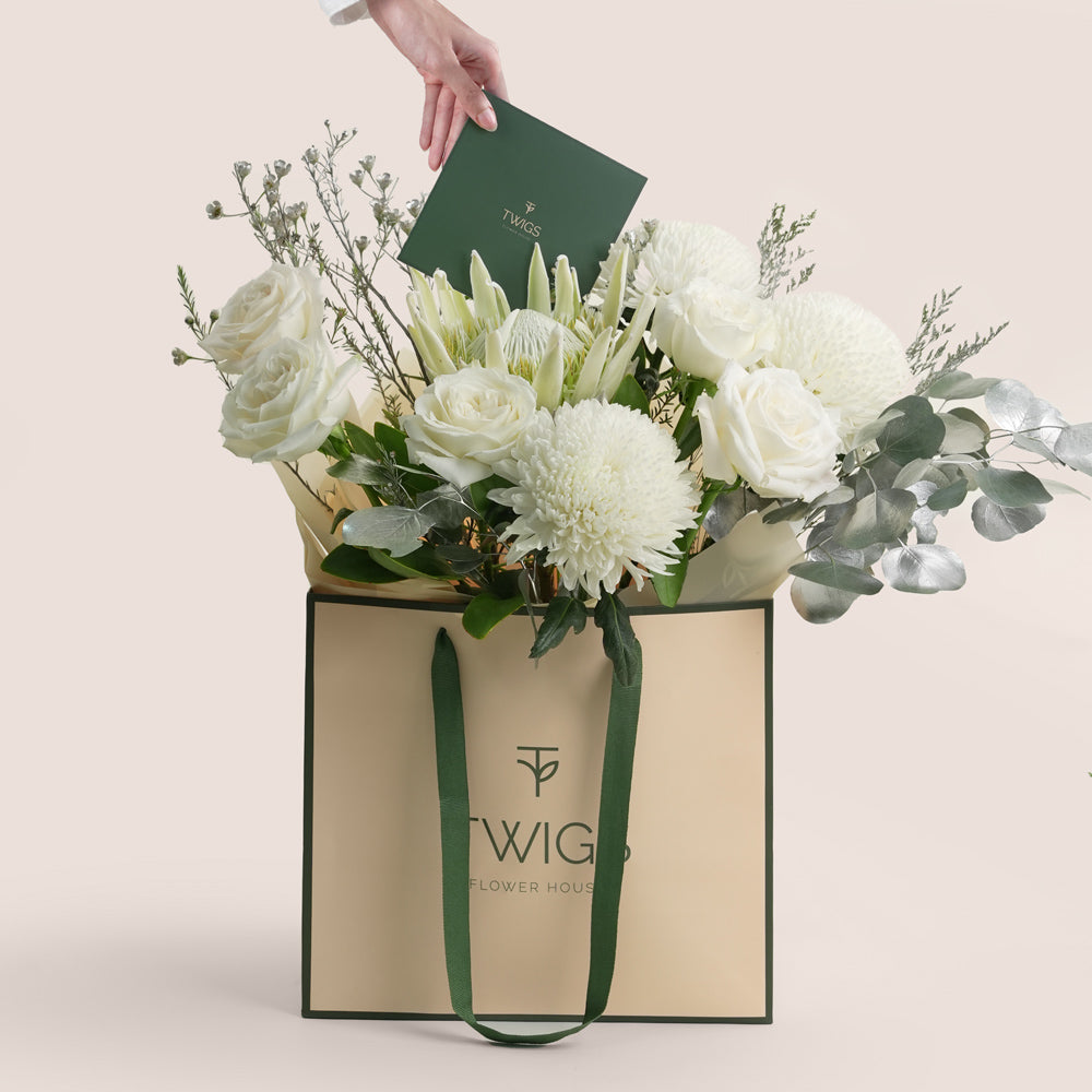 Floral arrangement in a Twigs Flower House bag with a hand holding a card on a light pink background