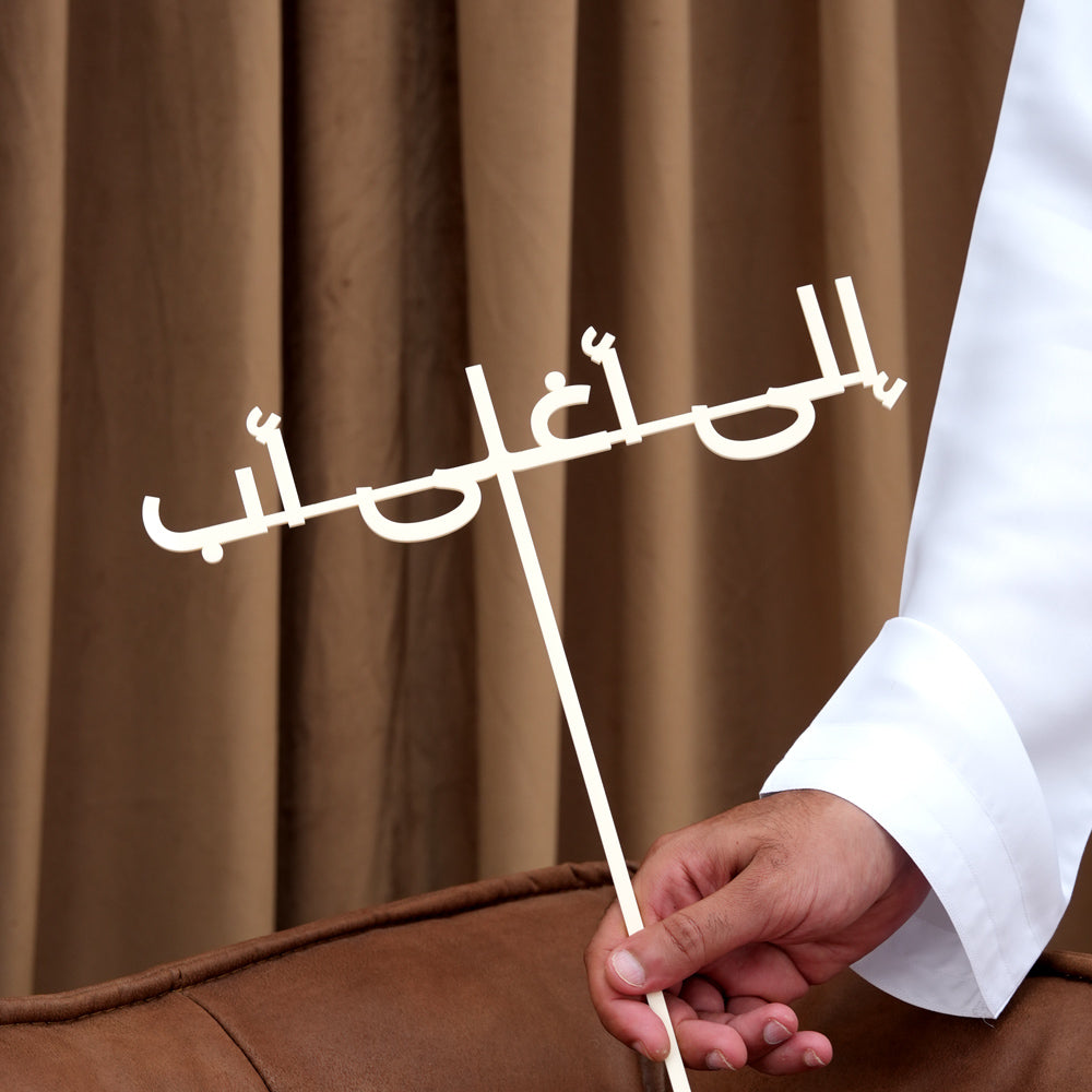Father's Day Arabic message topper reading إلى أغلى بابا held against a brown curtain background, TWIGS flower arrangement add-on Dubai