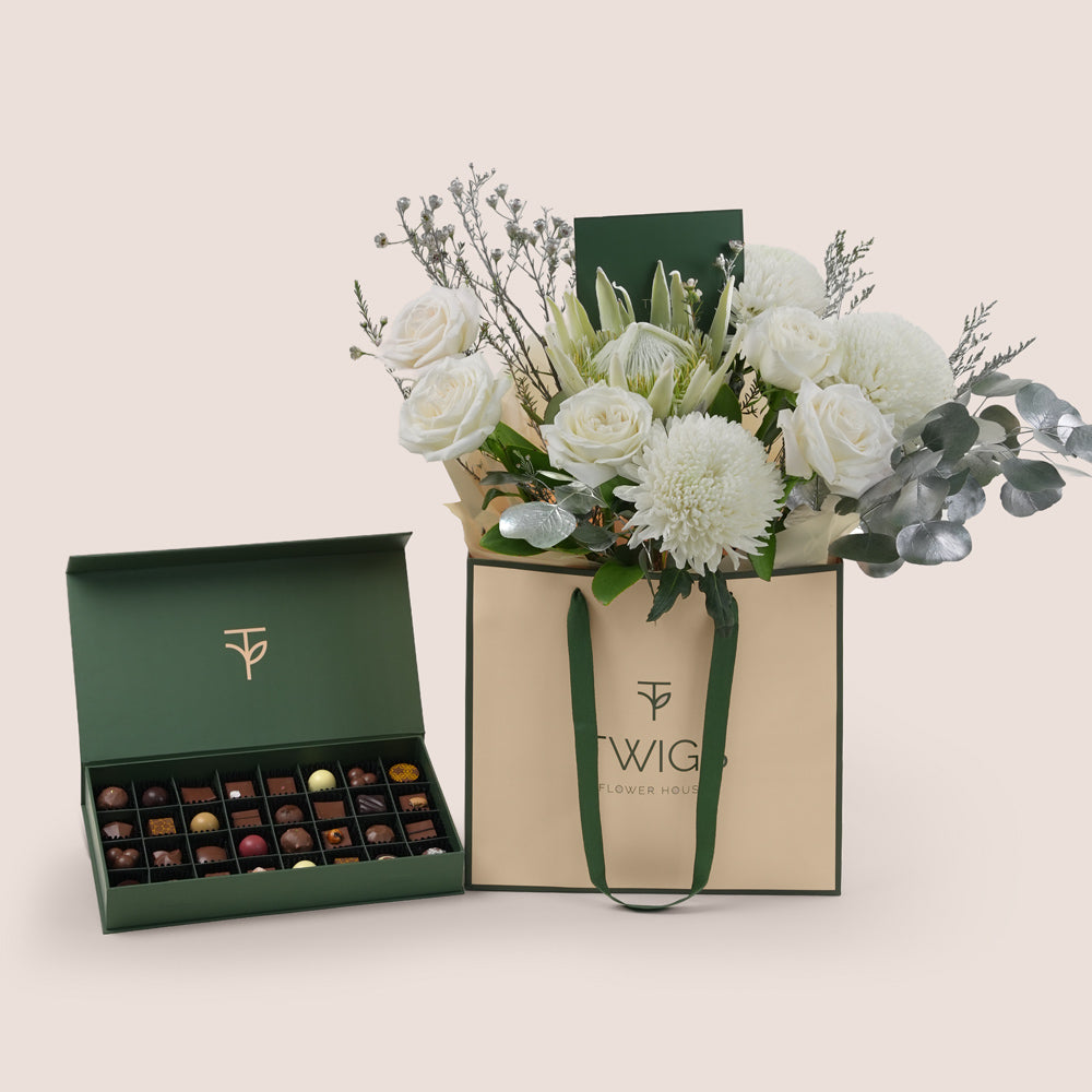 A luxury gift set from Twigs Flower House with a bouquet of white roses, white chrysanthemums, and silver-sprayed eucalyptus in a beige and green branded bag, next to an open deep green box of assorted gourmet chocolates.