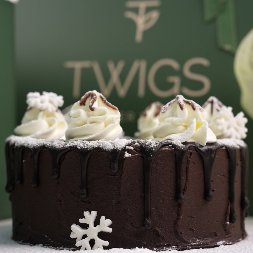 A New Year gift set from Twigs Flower House featuring white flowers (chrysanthemums and anthuriums), a small chocolate cake, a deep green gift box, a dark green carrier bag with the Twigs logo, and a bouquet of white and silver helium balloons