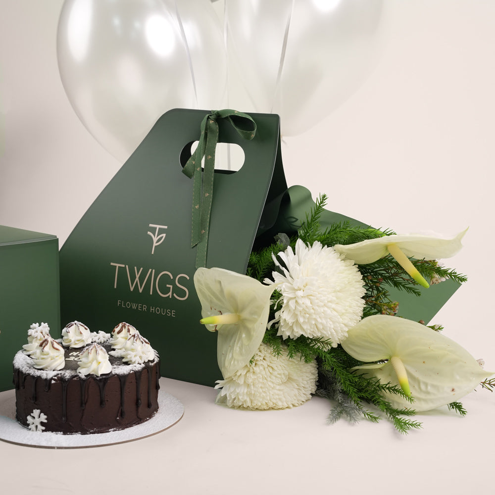 A New Year gift set from Twigs Flower House featuring white flowers (chrysanthemums and anthuriums), a small chocolate cake, a deep green gift box, a dark green carrier bag with the Twigs logo, and a bouquet of white and silver helium balloons