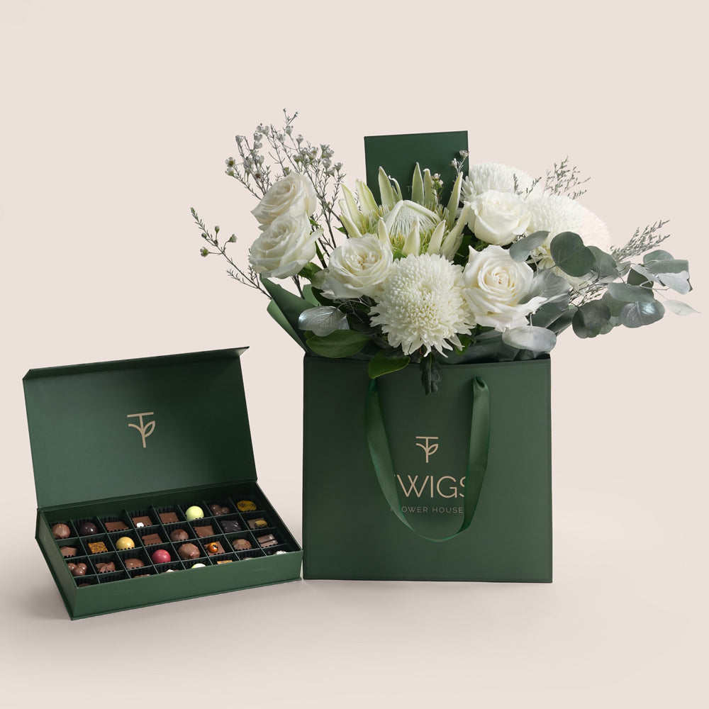 A luxury gift set from Twigs Flower House with a bouquet of white roses, white chrysanthemums, and silver-sprayed eucalyptus in a beige and green branded bag, next to an open deep green box of assorted gourmet chocolates.