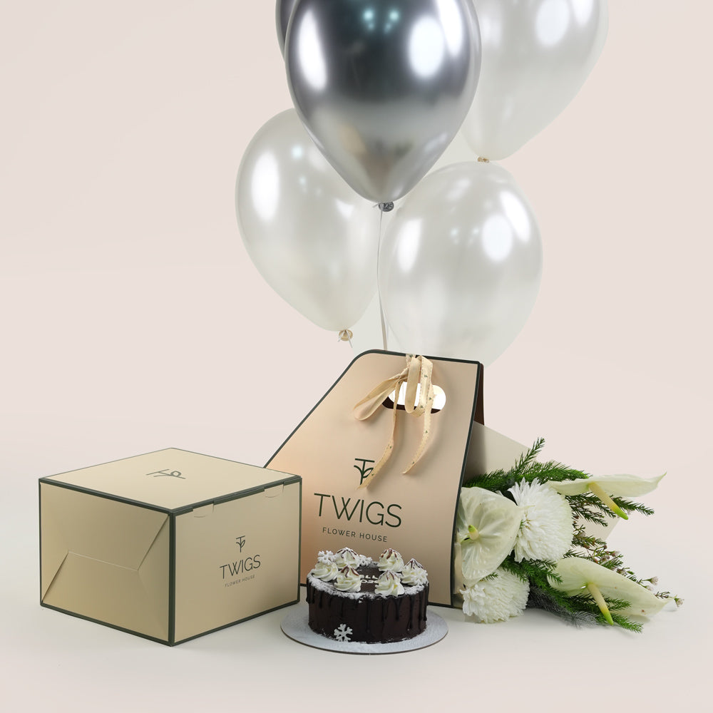 A New Year gift set from Twigs Flower House featuring white flowers (chrysanthemums and anthuriums), a small chocolate cake, a deep green gift box, a beige carrier bag with the Twigs , and a bouquet of white and silver helium balloons