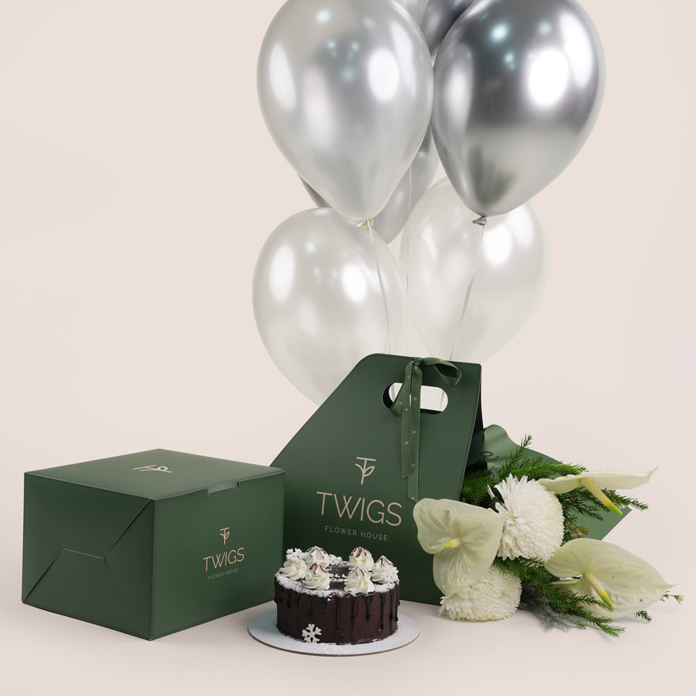 A New Year gift set from Twigs Flower House featuring white flowers (chrysanthemums and anthuriums), a small chocolate cake, a deep green gift box, a dark green carrier bag with the Twigs , and a bouquet of white and silver helium balloons