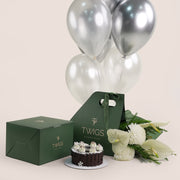 A New Year gift set from Twigs Flower House featuring white flowers (chrysanthemums and anthuriums), a small chocolate cake, a deep green gift box, a dark green carrier bag with the Twigs , and a bouquet of white and silver helium balloons