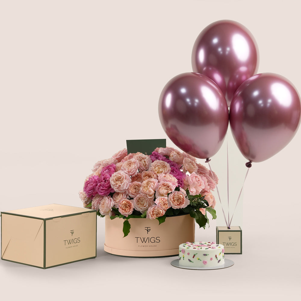 Grande round flower box with peach Juliet spray roses and cerise accents, paired with a 1kg birthday cake and three chrome dark pink balloons in TWIGS beige or green packaging, available for same day flower delivery in Dubai when you buy flowers online.