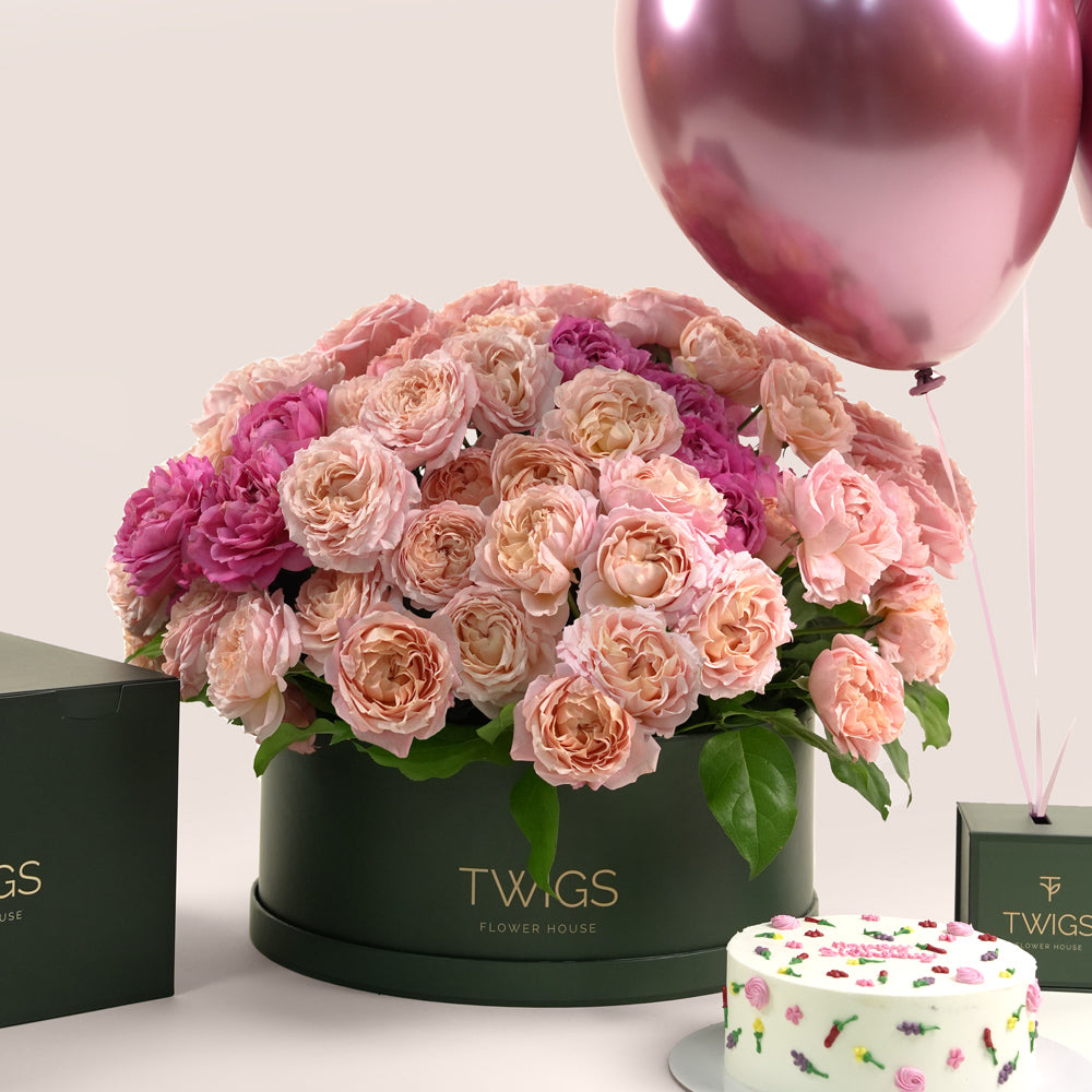 Grande round flower box with peach Juliet spray roses and cerise accents, paired with a 1kg birthday cake and three chrome dark pink balloons in TWIGS beige or green packaging, available for same day flower delivery in Dubai when you buy flowers online.