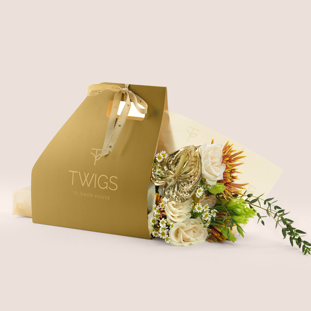 Aurelia, a TWIGS Gold gift bag with garden roses, chamomiles, saffina mums and a gold painted anthurium leaf accent, on a light beige background.