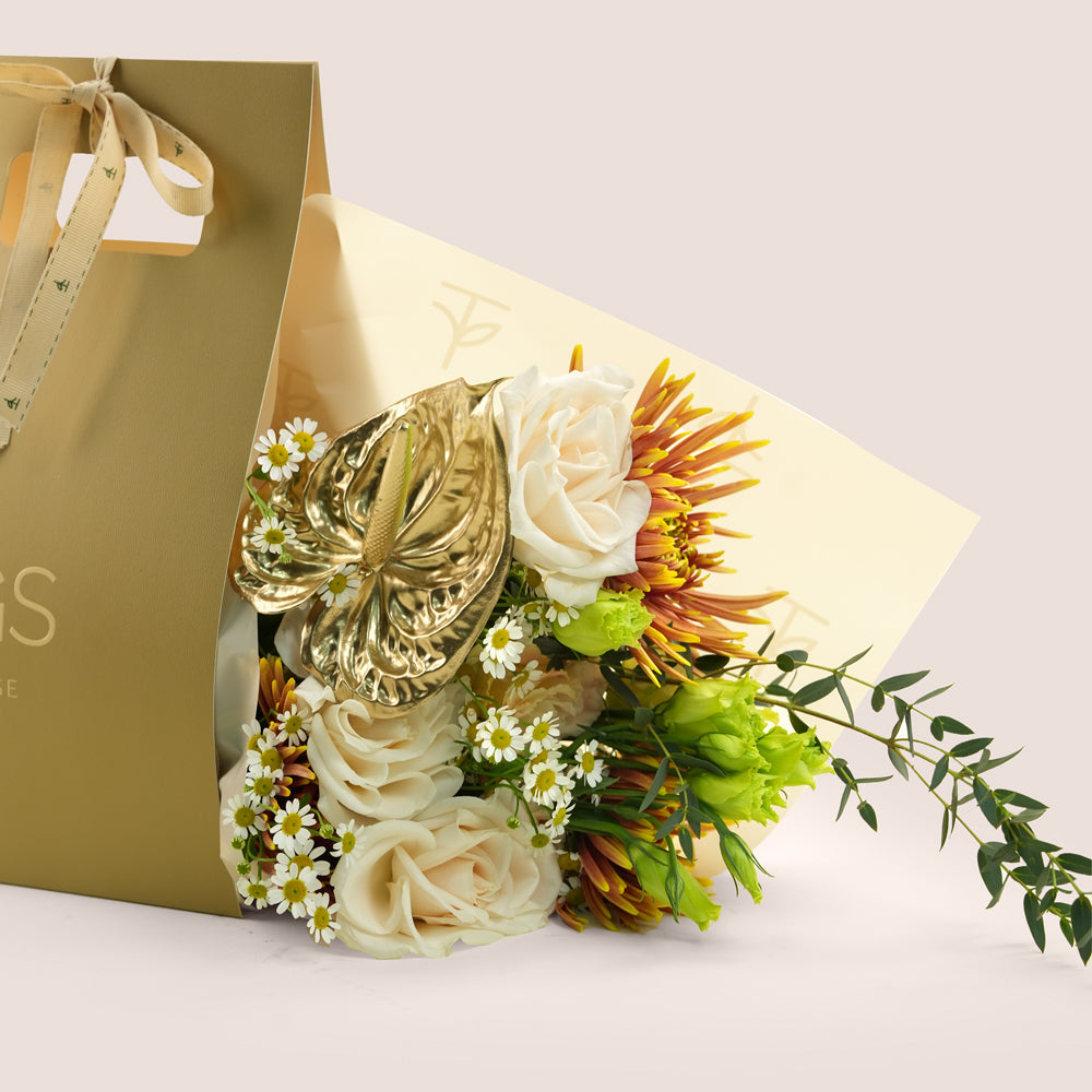 Aurelia, a TWIGS Gold gift bag with garden roses, chamomiles, saffina mums and a gold painted anthurium leaf accent, on a light beige background.