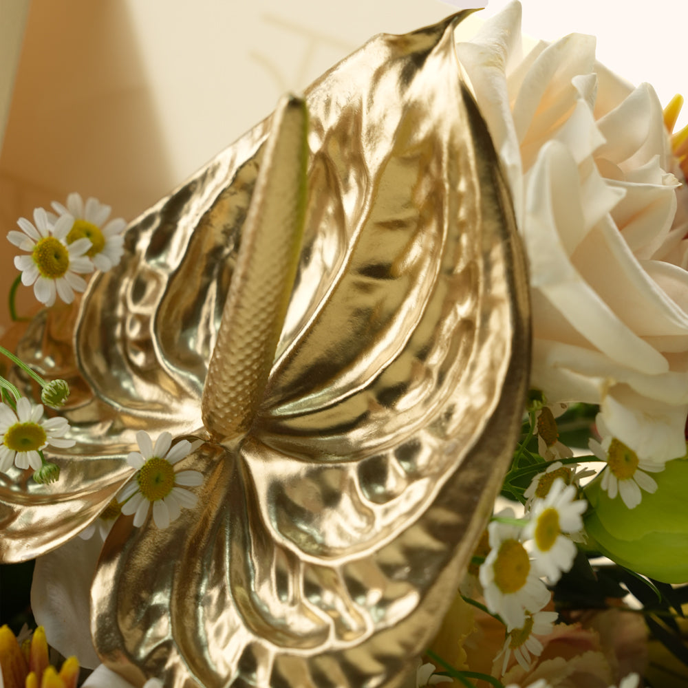A close-up of a metallic gold anthurium flower arranged with white daisies and cream roses in a floral setting.