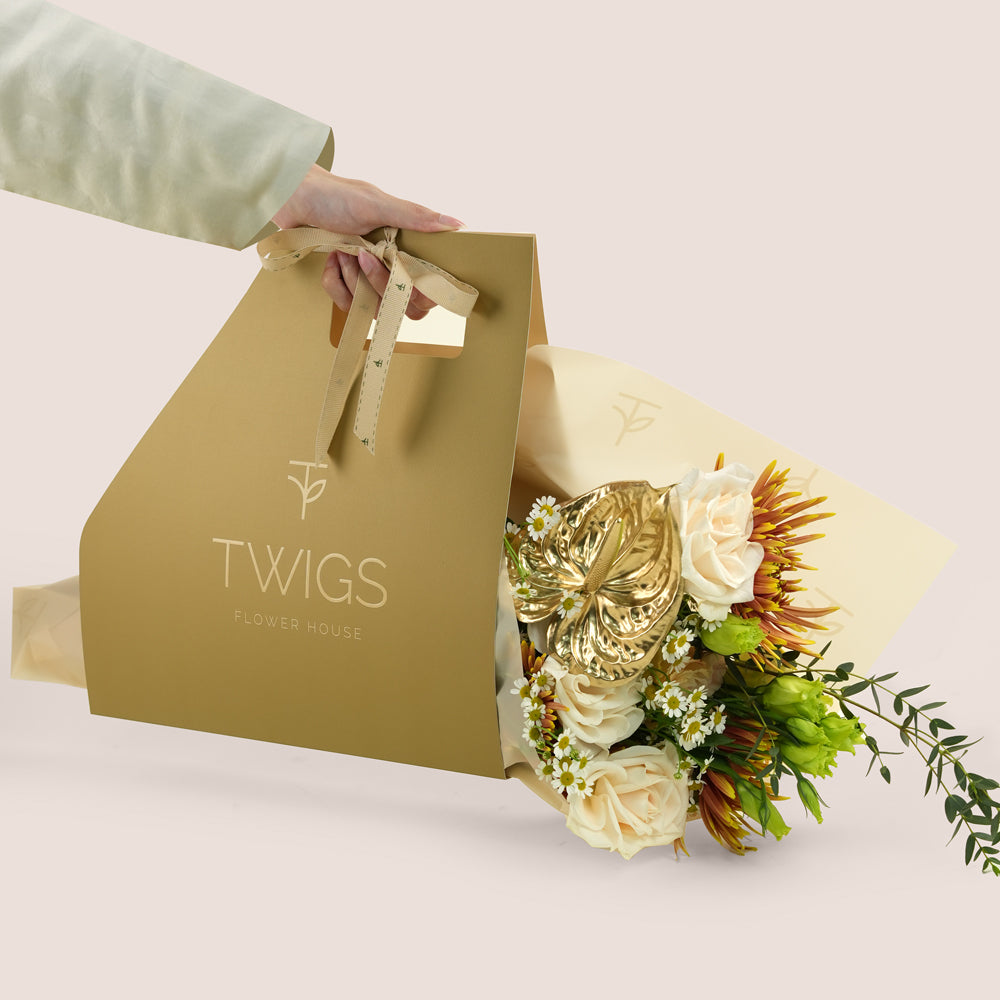 Aurelia, a TWIGS Gold gift bag with garden roses, chamomiles, saffina mums and a gold painted anthurium leaf accent, on a light beige background.