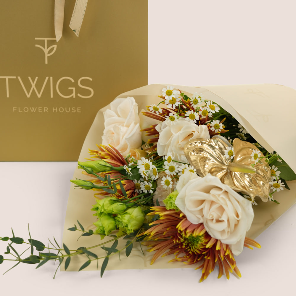 A floral bouquet wrapped in beige paper, featuring cream roses, small white daisy-like flowers, green buds, and orange-brown chrysanthemum-style blooms, with a metallic gold decorative accent. A gold TWIGS FLOWER HOUSE gift bag is visible behind the bouquet on a clean off-white background.