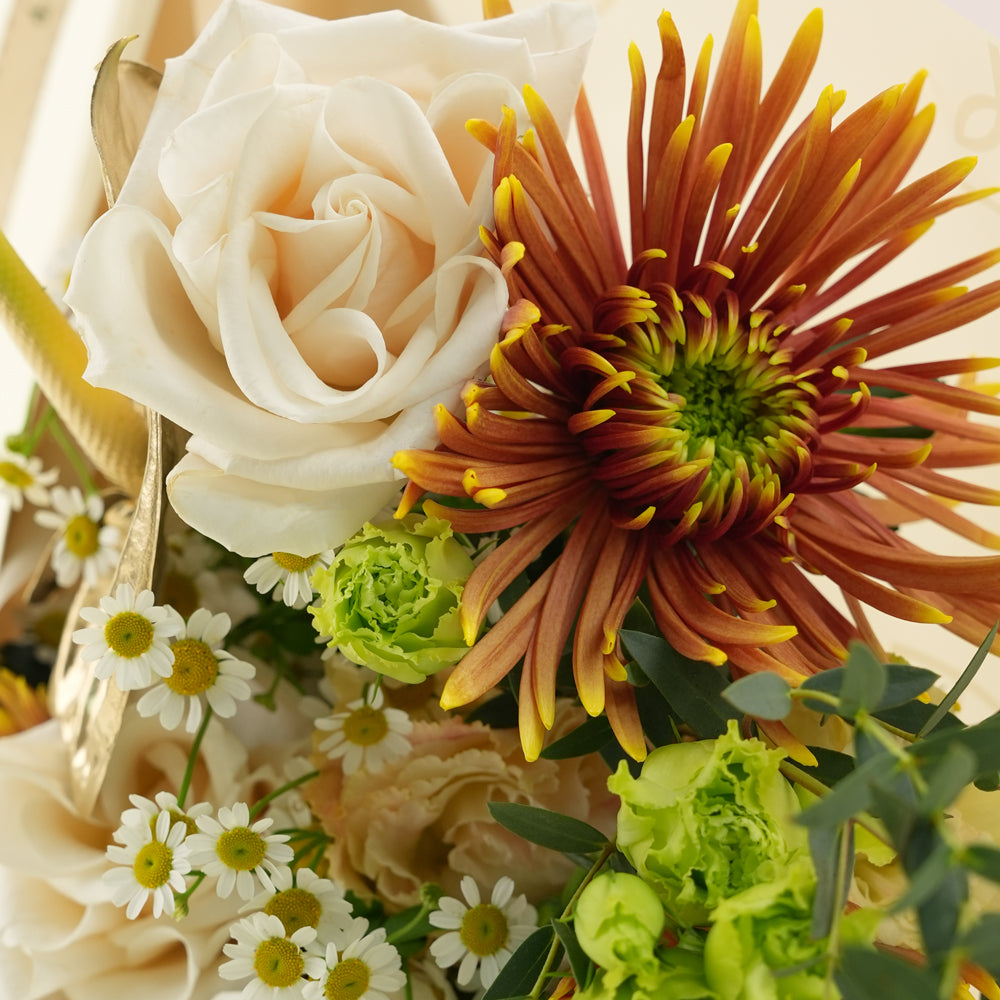 A close-up floral arrangement featuring a large cream rose, a prominent orange-brown daisy-like flower with yellow tips, small white daisies, pale green flowers, and green foliage on a warm cream background.