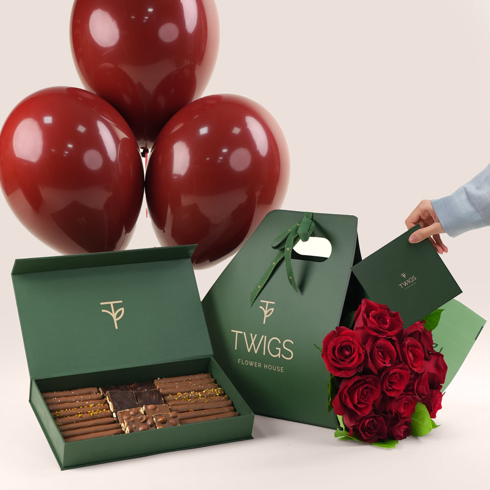Green gift box with chocolates, red balloons, and a bouquet of red roses on a light background.