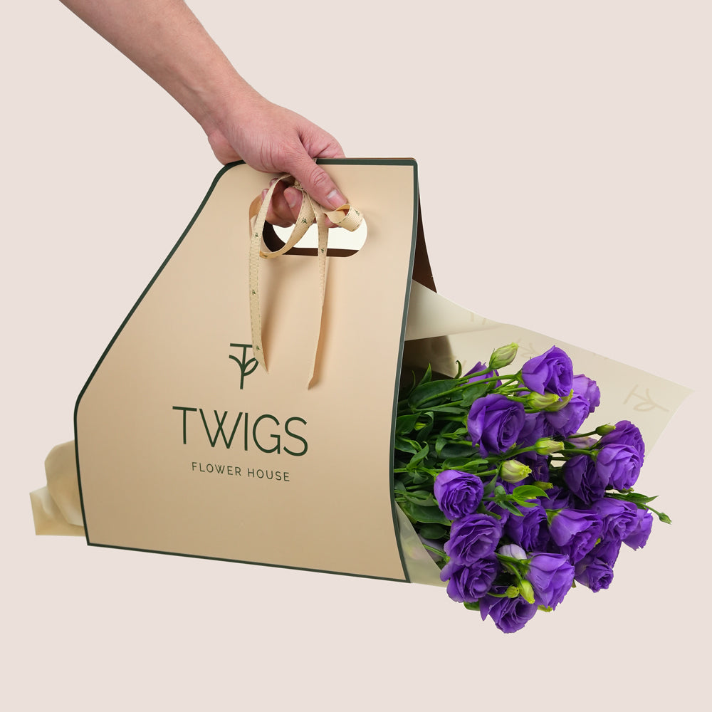 Hand holding a beige 'TWIGS Flower House' bag with a bouquet of purple flowers on a light background