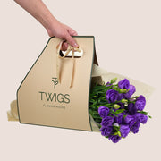 Hand holding a beige 'TWIGS Flower House' bag with a bouquet of purple flowers on a light background