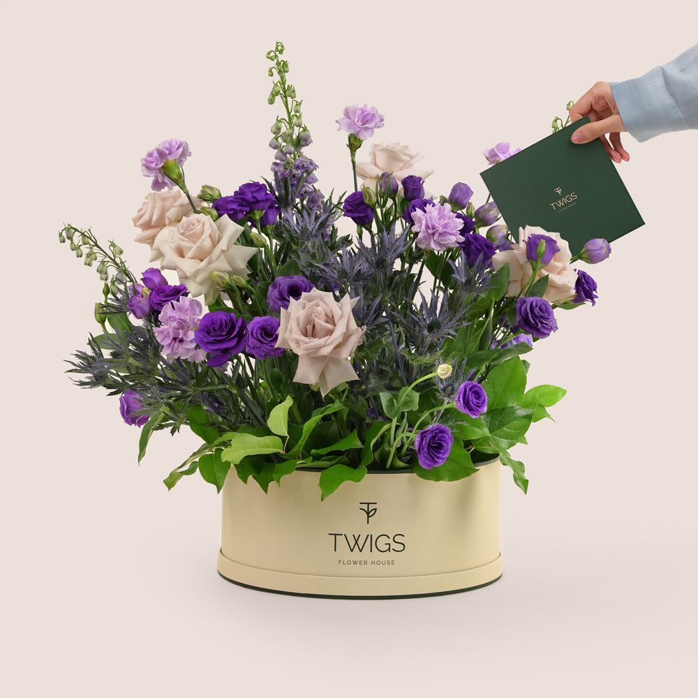 Grande round flower box with quicksand roses, purple lisianthus, carnations, delphiniums, and eryngium in TWIGS beige or green packaging, available for same day flower delivery in Dubai when you buy flowers online.