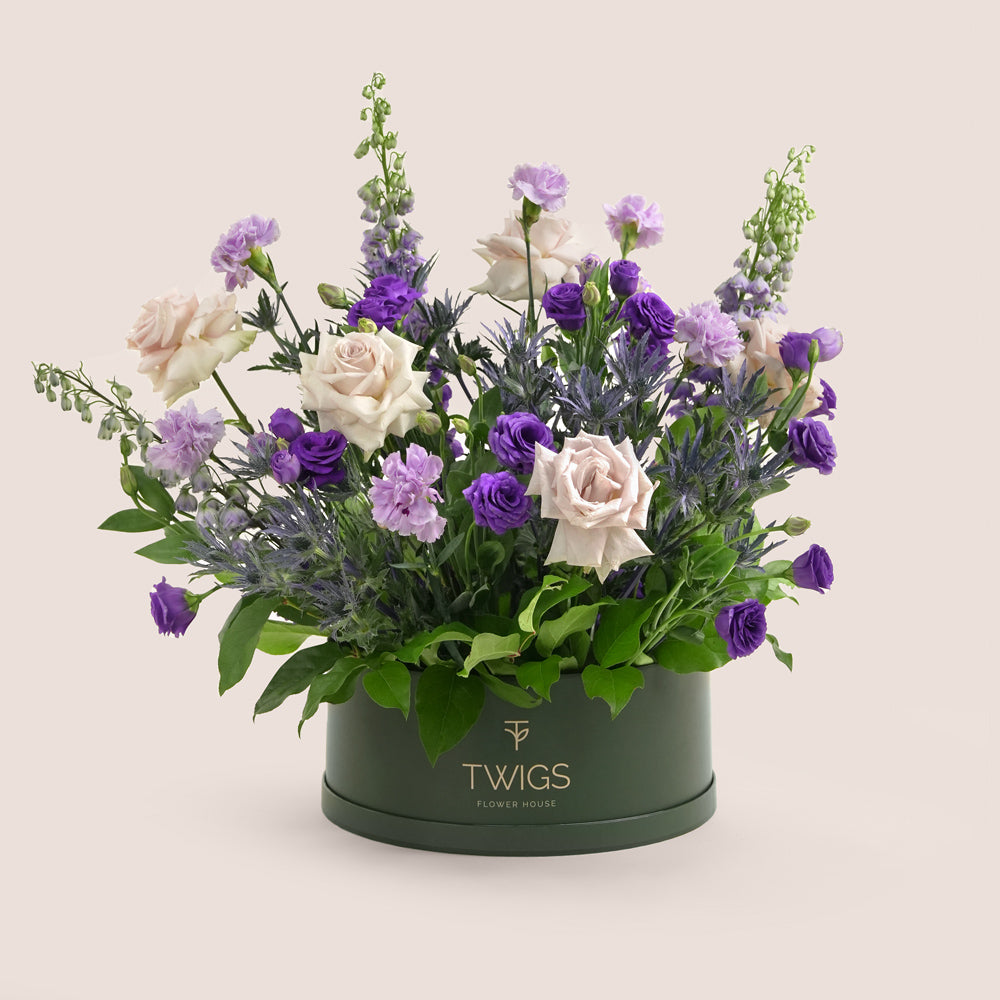 Grande round flower box with quicksand roses, purple lisianthus, carnations, delphiniums, and eryngium in TWIGS beige or green packaging, available for same day flower delivery in Dubai when you buy flowers online.
