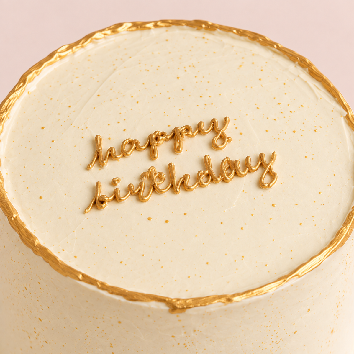 A round 1kg TWIGS Gold celebration cake with gold trim and a "Happy Birthday" message piped in gold icing, held on a white cake stand.