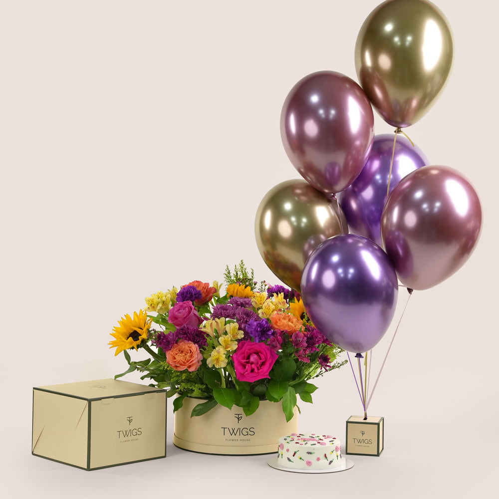 Festive grande round flower box with sunflowers, roses, alstroemerias, chrysanthemums, and wax flowers, paired with a 1kg birthday cake and six chrome balloons in TWIGS beige or green packaging, available for same day flower delivery in Dubai when you buy flowers online.
