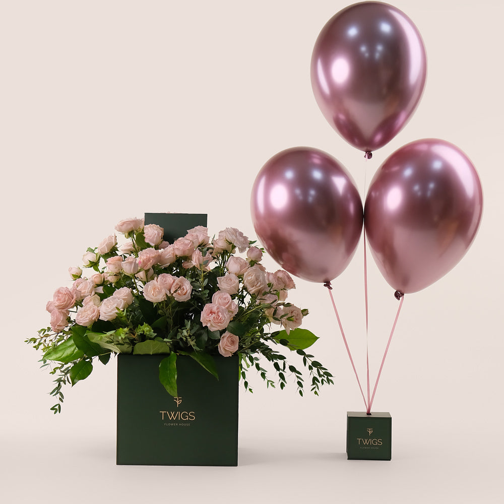 A signature square flower box with a bunch of soft pink bombastic roses and two metallic pink balloons tied to it, with the TWIGS logo on the box.