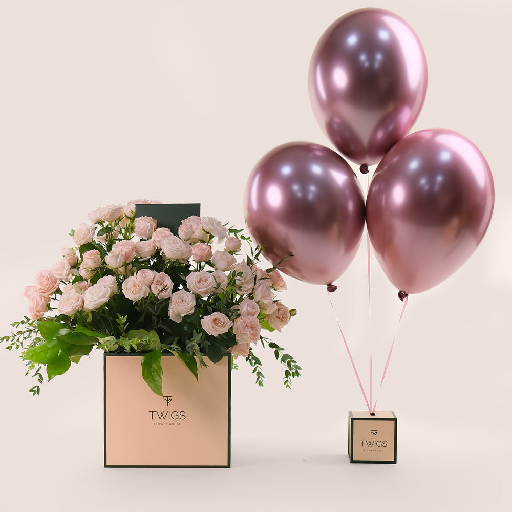 A signature square flower box with a arrangement of soft pink bombastic roses and metallic pink balloons tied to it, with the TWIGS logo on the box.