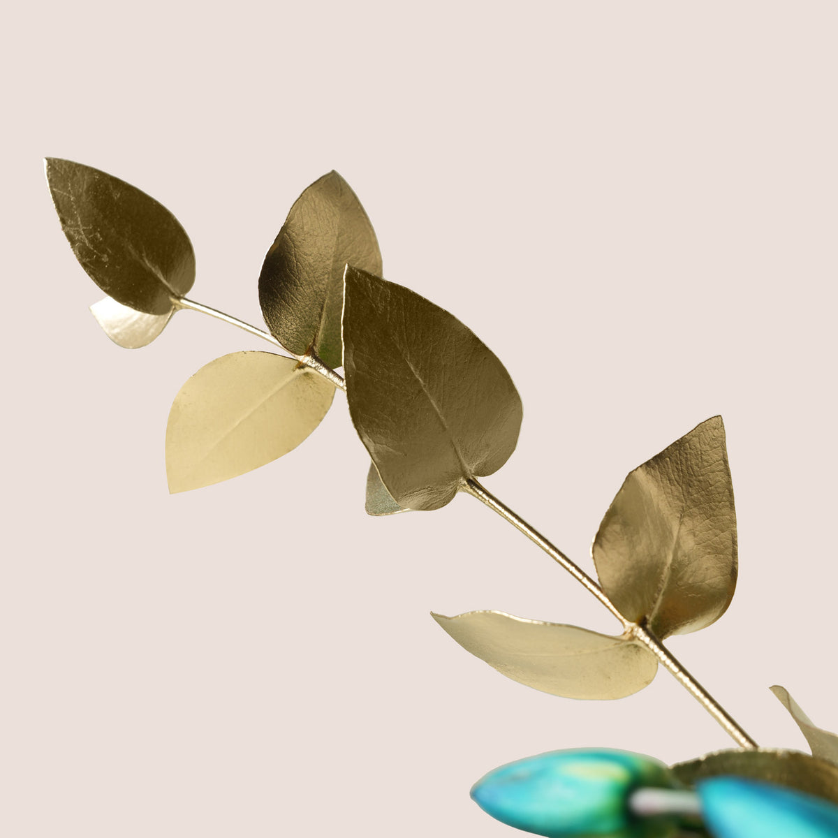 A decorative branch with metallic gold-toned leaves on a thin stem, photographed against a plain warm beige background. A small portion of a blue-green object is visible at the bottom edge.