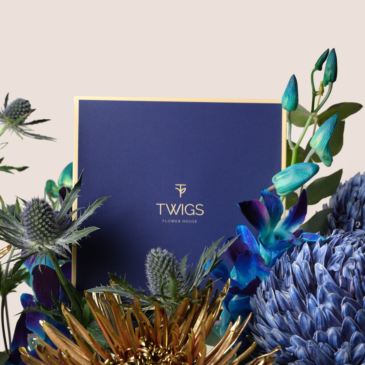 A navy blue floral arrangement box with gold foil branding reading 'TWIGS FLOWER HOUSE' surrounded by decorative flowers and greenery on a light beige background.