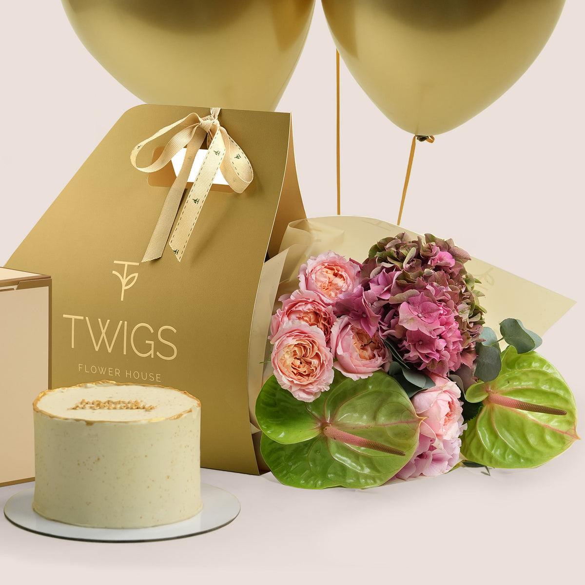 Isadora Celebration, flowers and cake gift set: a TWIGS Gold carry bag with pink roses, pink hydrangeas, and green anthurium leaves, paired with a 1kg cake and three gold chrome balloons.