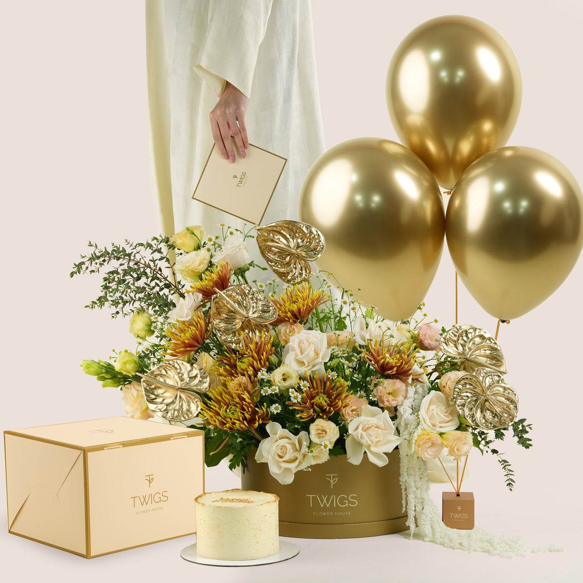 Aurelia Celebration, a luxury TWIGS Gold Garden box arrangement of white and peach roses, chrysanthemums, and gold anthurium leaves, paired with a 1kg cake and three gold chrome balloons.