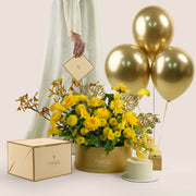 Solara Celebration, a vibrant yellow floral arrangement of calla lilies, chrysanthemums, roses, and gold anthurium leaves in a round TWIGS Gold box, paired with a 1kg cake and three gold chrome balloons.