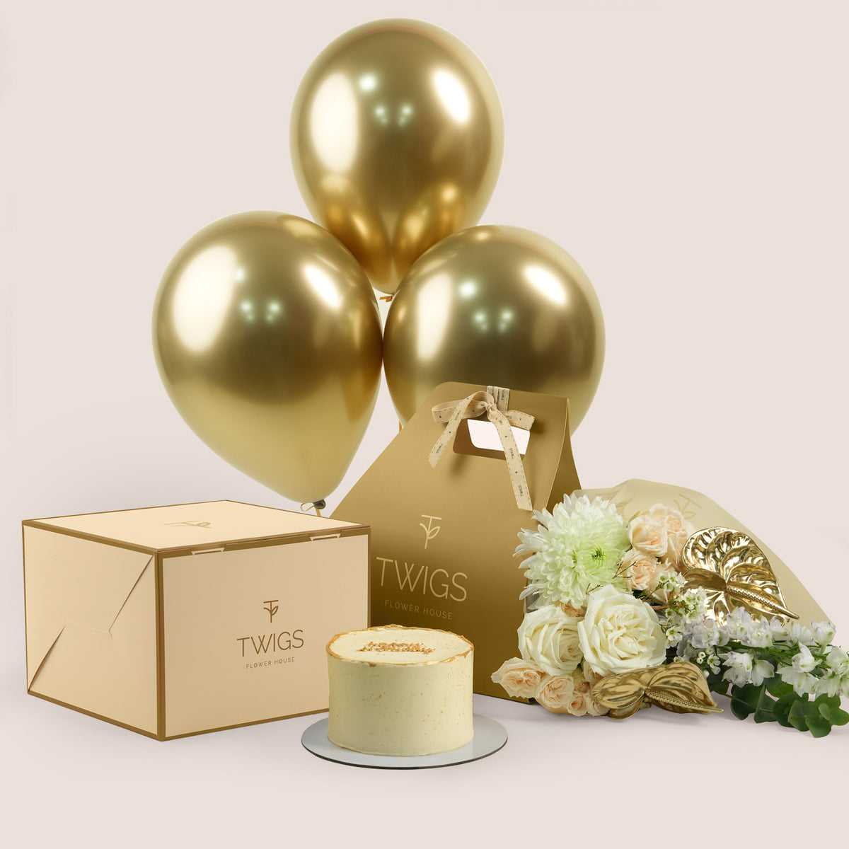 Lumina Celebration, flowers and cake with balloons gift set: a TWIGS Gold carry bag with white roses, peach spray roses, chrysanthemums, and a gold anthurium leaf accent, paired with a 1kg cake and three gold chrome balloons.