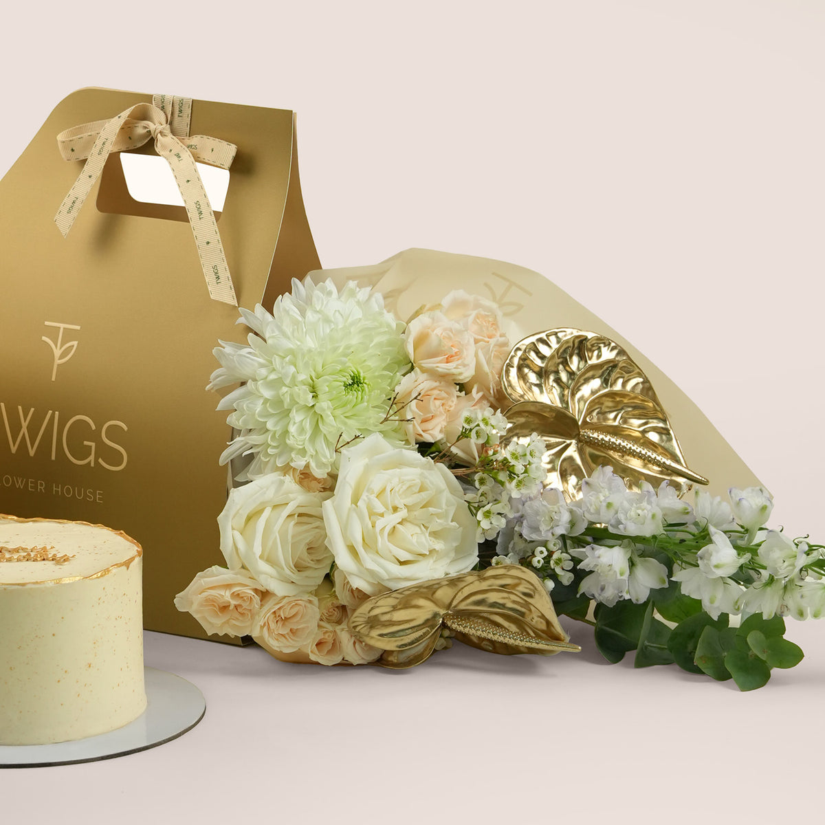 Lumina Celebration, flowers and cake with balloons gift set: a TWIGS Gold carry bag with white roses, peach spray roses, chrysanthemums, and a gold anthurium leaf accent, paired with a 1kg cake and three gold chrome balloons.