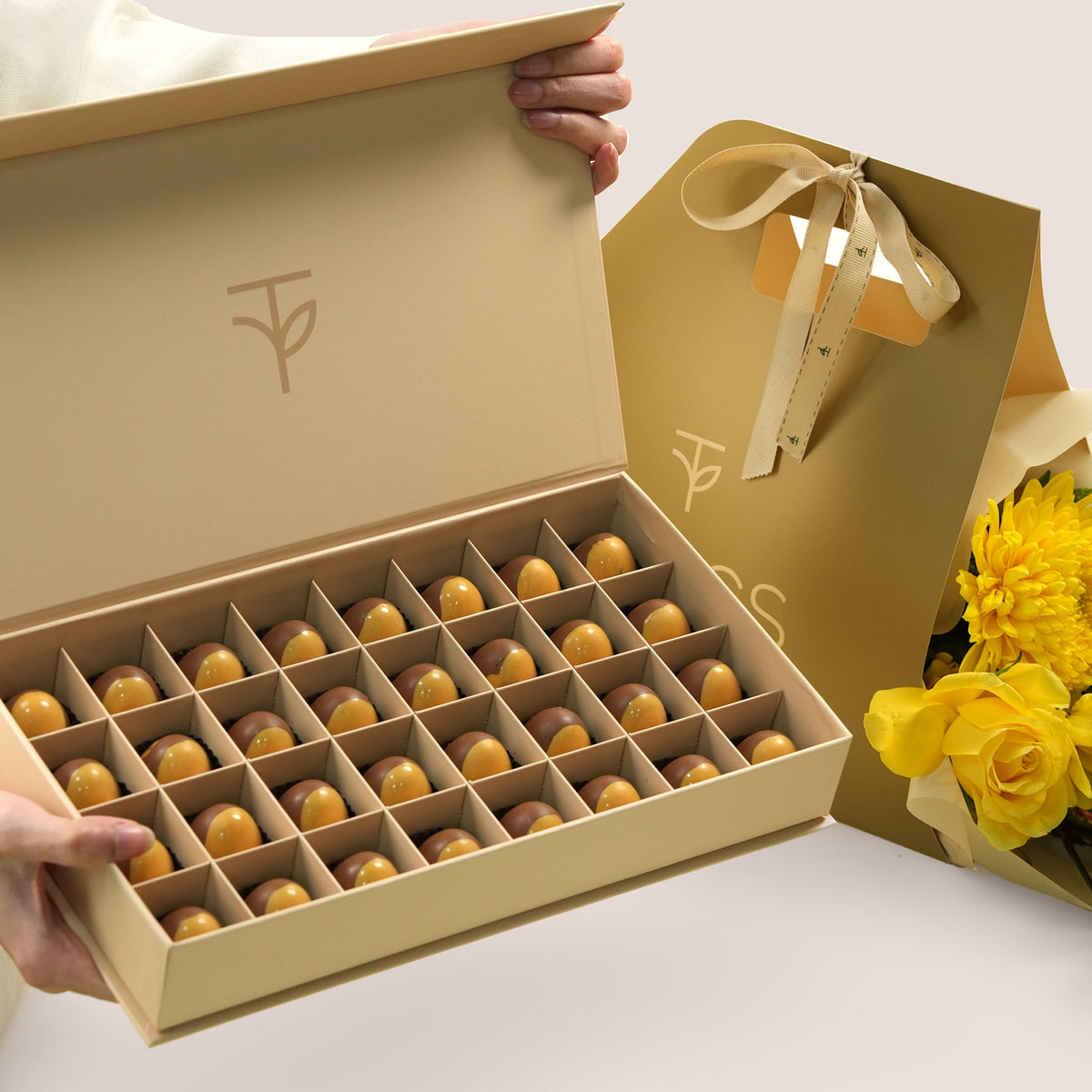 Solara Pairing, flowers and chocolate gift set: a TWIGS Gold carry bag with yellow roses, chrysanthemums, and a gold anthurium leaf accent, paired with a 32 piece premium bonbon box.