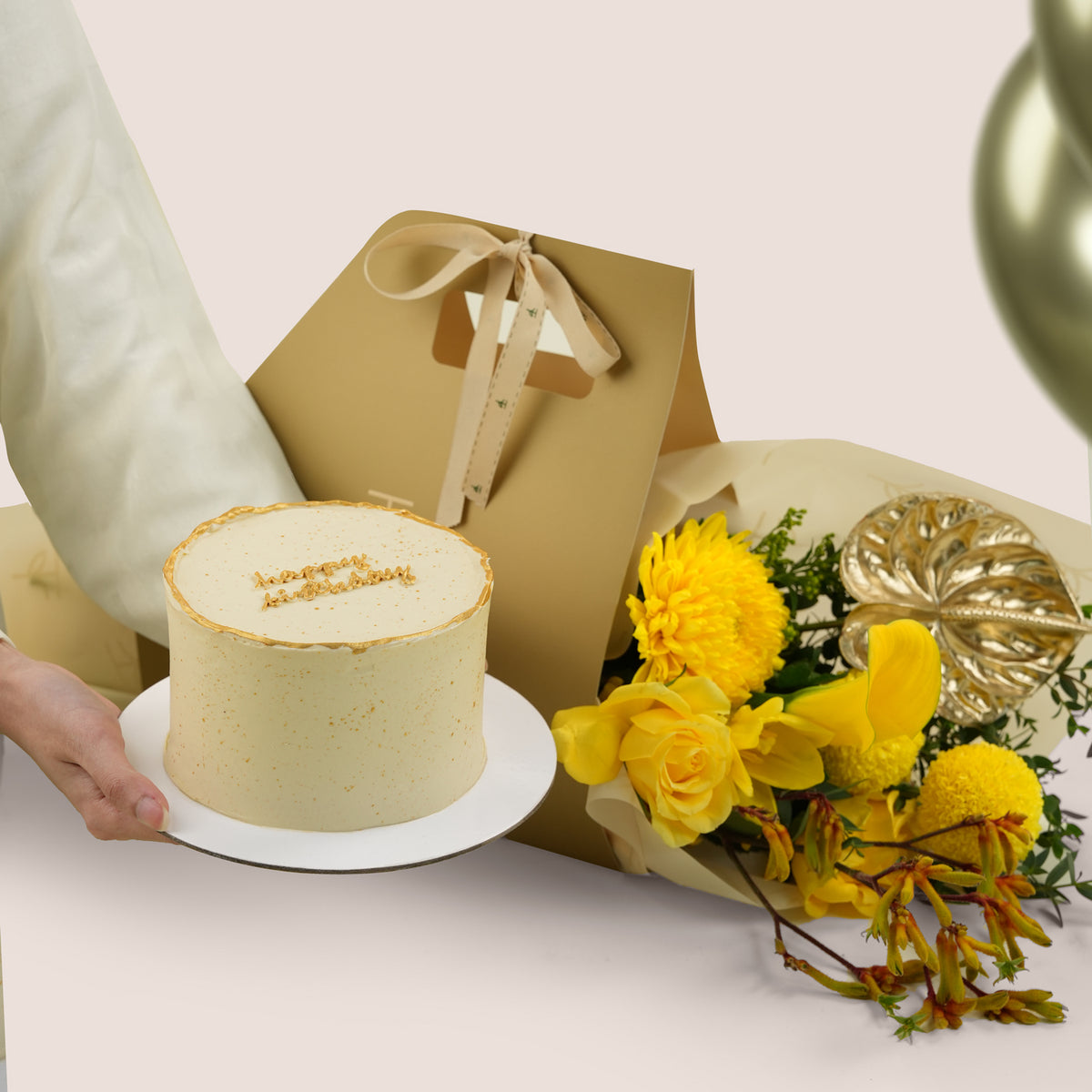 Solara Celebration, a TWIGS Gold carry bag with yellow roses, chrysanthemums, kangaroo paw, and a gold anthurium leaf accent, paired with a 1kg cake and three gold chrome balloons.