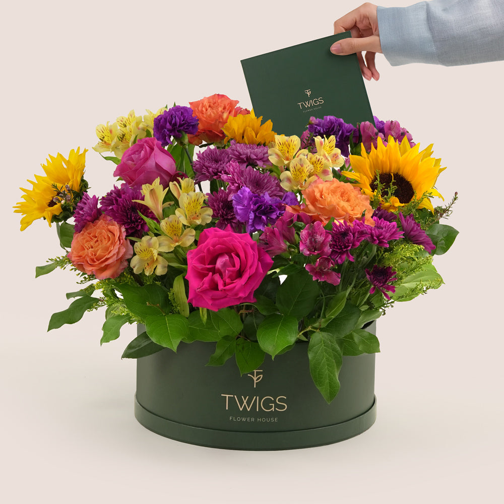 Festive grande round flower box with sunflowers, roses, alstroemerias, chrysanthemums, and wax flowers in TWIGS beige or green packaging, available for same day flower delivery in Dubai when you buy flowers online.