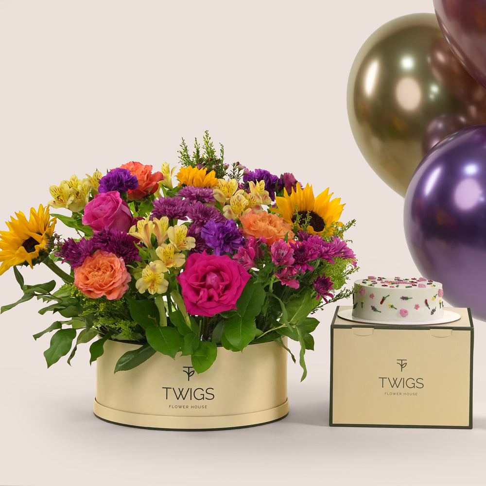 Festive grande round flower box with sunflowers, roses, alstroemerias, chrysanthemums, and wax flowers, paired with a 1kg birthday cake and six chrome balloons in TWIGS beige or green packaging, available for same day flower delivery in Dubai when you buy flowers online.