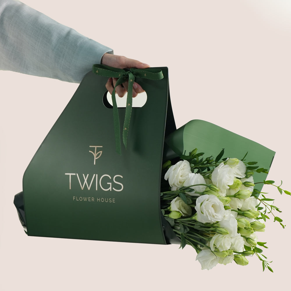 Green bunch with 'TWIGS FLOWER HOUSE' branding holding a bouquet of white flowers on a light beige background.